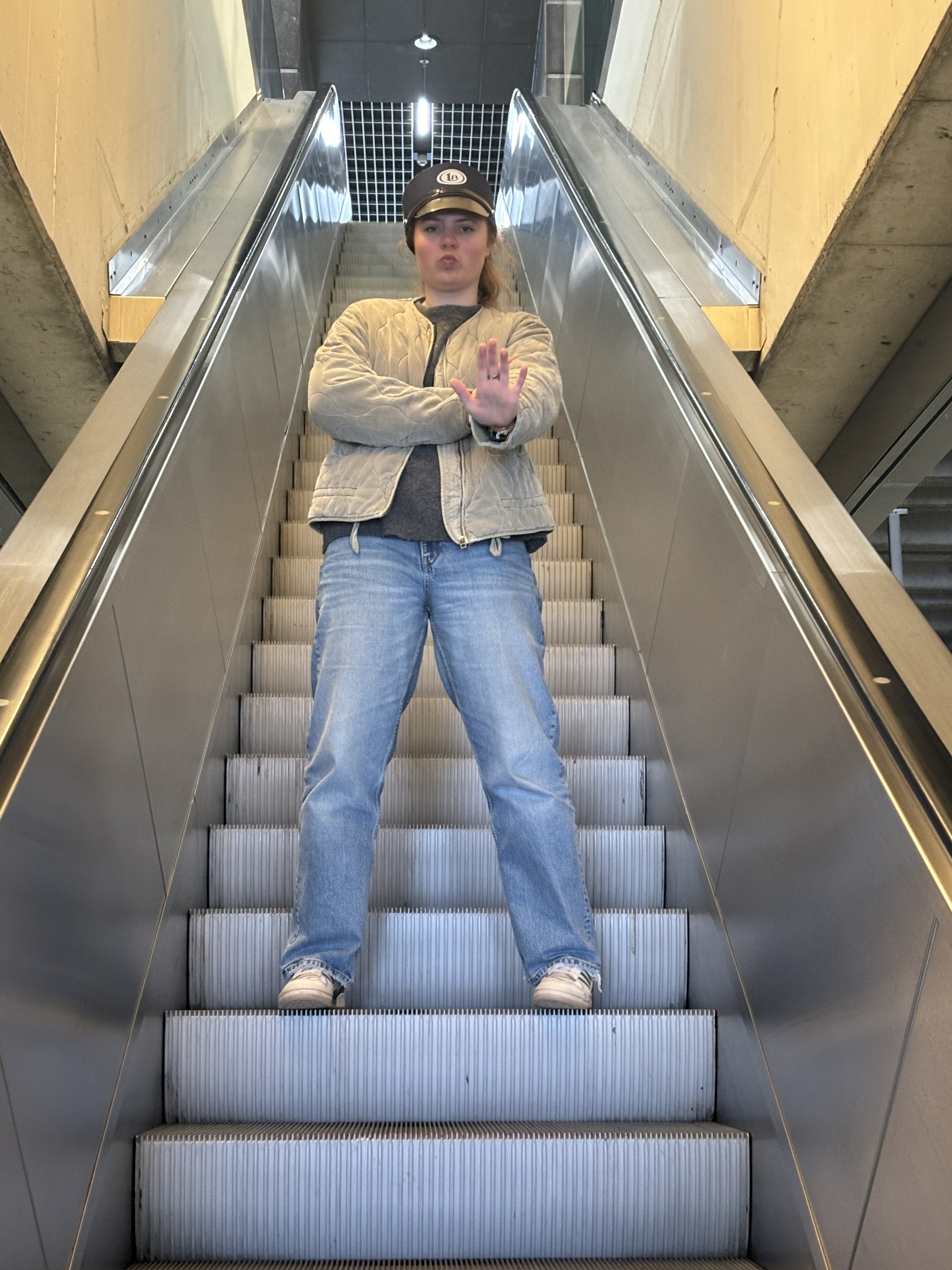 Photo d'Alice dans les escalators. Elle porte un képi avec le logo Late et croise les bras, une main en avant.