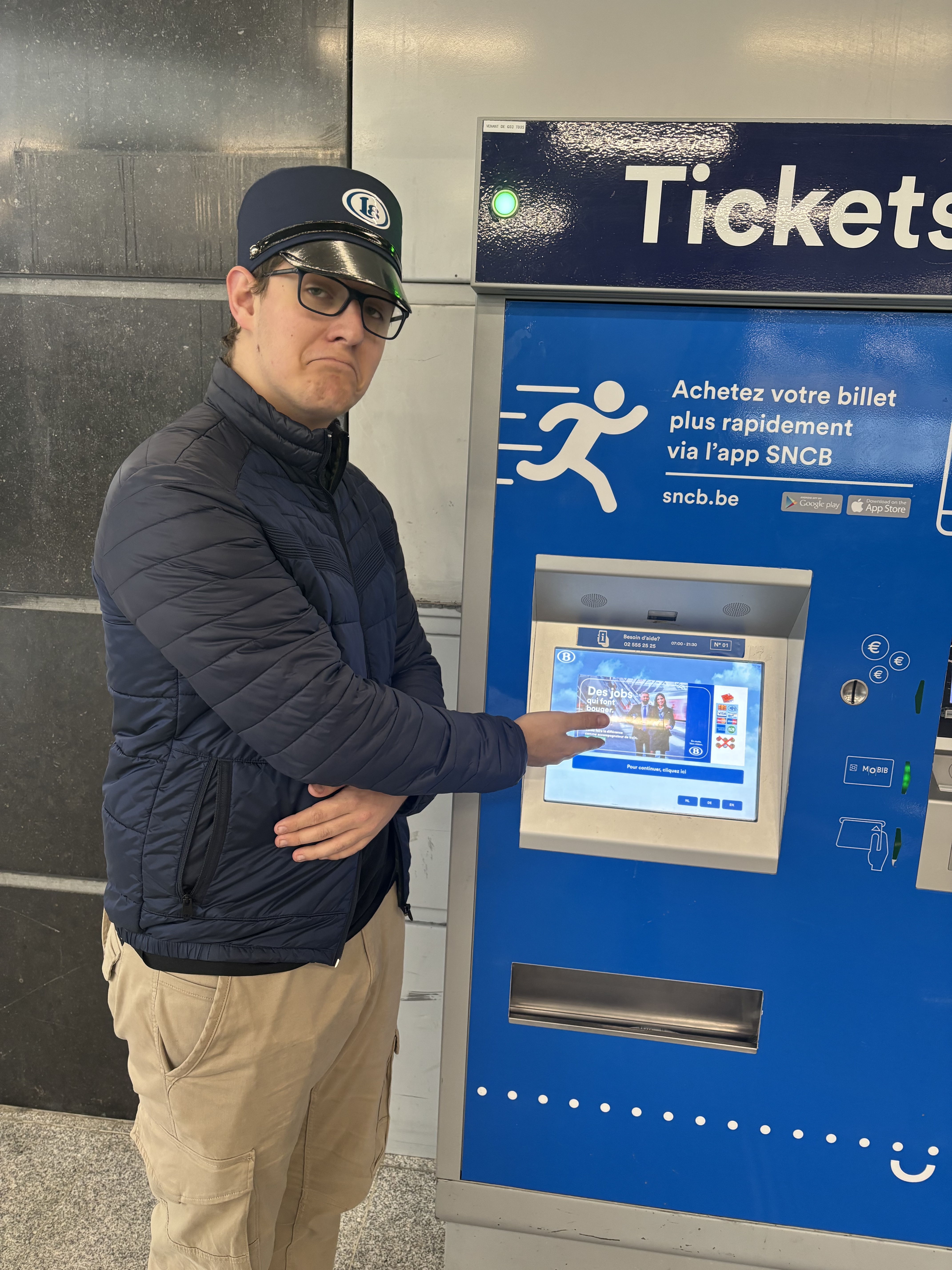 Photo de Guillaume présentant une borne permetant de prendre des billets de train. Il porte un képi Late.