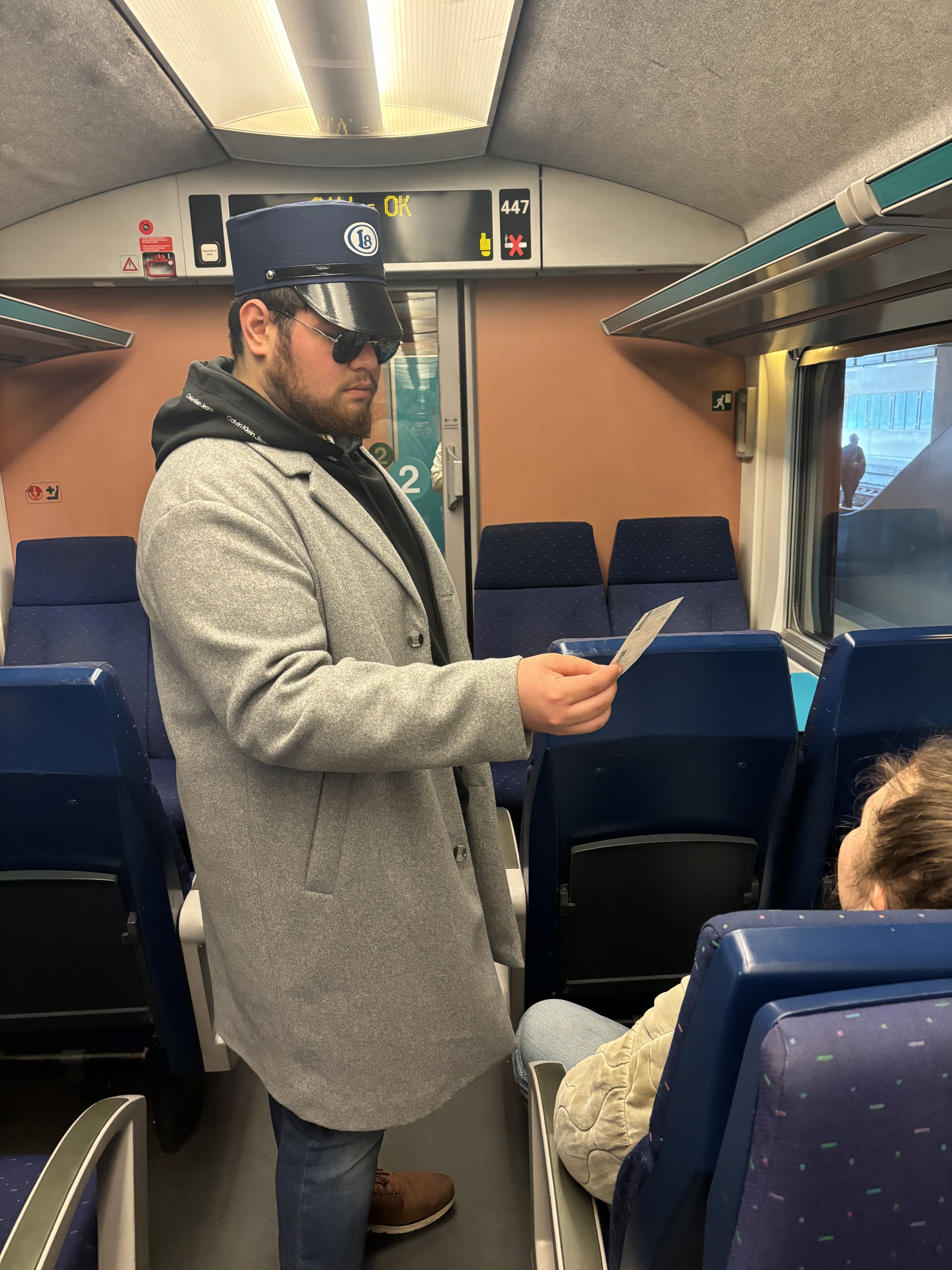 photo de Maxime, controlant un voyageur dans un train. Il porte un képi avec le logo Late et des lunettes de soleil. Il inspecte le billet du voyageur. 